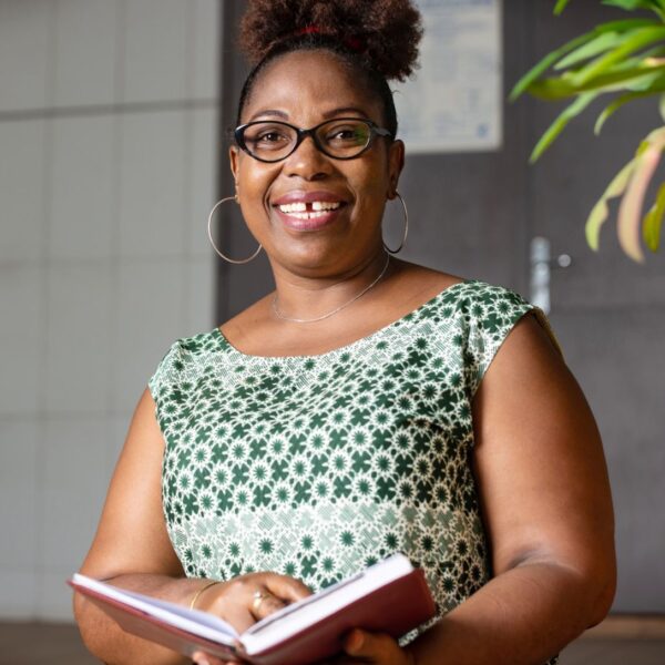 A middle-aged woman with coffee coloured skin smiles at the camera. Her black, tightly coiled hair is tied up at the top of her head. She wears black rimmed glasses over her dark brown eyes, large, gold hoop earrings and a white and green geometric patterned sleeveless dress. She holds an open hardback book in her hands.