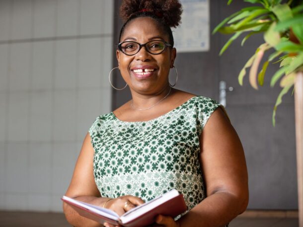 A middle-aged woman with coffee coloured skin smiles at the camera. Her black, tightly coiled hair is tied up at the top of her head. She wears black rimmed glasses over her dark brown eyes, large, gold hoop earrings and a white and green geometric patterned sleeveless dress. She holds an open hardback book in her hands.