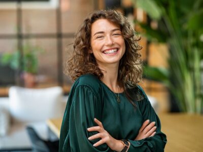 A young woman looks side-on at the camera and laughs. The pale freckled skin around her brown eyes creases with the laughter. She wears her brown, curly, shoulder-length hair down and an emerald green blouse.