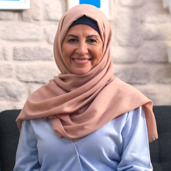An older lady sits on a sofa and smiles at the camera. Her olive skin creases with smile lines around her dark brown eyes. She is wearing a pale rose coloured headscarf and a light blue blouse.
