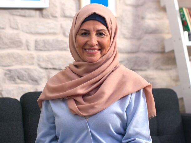 An older lady sits on a sofa and smiles at the camera. Her olive skin creases with smile lines around her dark brown eyes. She is wearing a pale rose coloured headscarf and a light blue blouse.