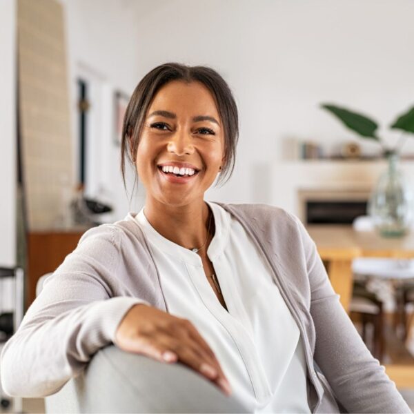 A woman sits facing sideways on a sofa as she laughs at the person behind the camera. Her dark brown hair is pulled back with a few loose sections falling over the caramel toned skin of her face. Her eyes are very dark brown and creased by laughter lines. She is wearing a white blouse underneath a grey cardigan.