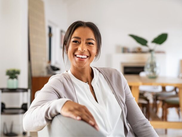 A woman sits facing sideways on a sofa as she laughs at the person behind the camera. Her dark brown hair is pulled back with a few loose sections falling over the caramel toned skin of her face. Her eyes are very dark brown and creased by laughter lines. She is wearing a white blouse underneath a grey cardigan.
