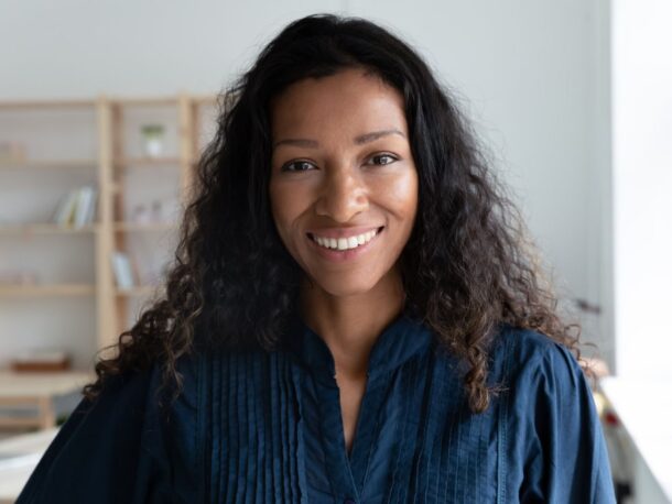 A woman with light coffee coloured skin and black shoulder length hair comprised of tight curls smiles at the camera. Her eyes are dark brown and she wears a dark blue collarless tunic.