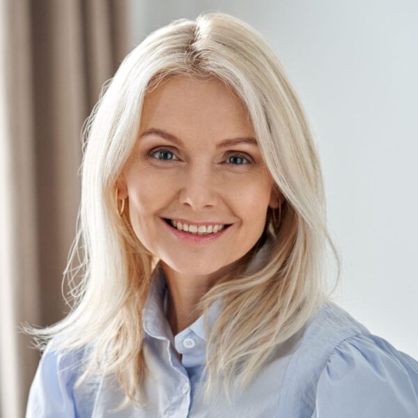 An older lady smiles at the camera, she is wearing subtle makeup, gold hoop earrings and a pale blue blouse. Her eyes are a dark blue and she wears her shoulder length golden white hair down.