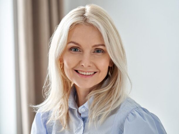 An older lady smiles at the camera, she is wearing subtle makeup, gold hoop earrings and a pale blue blouse. Her eyes are a dark blue and she wears her shoulder length golden white hair down.