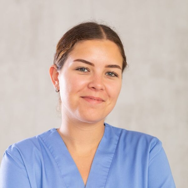 A young woman smiles at the camera. She's wearing blue scrubs and her brown hair is tied back. Her skin is lightly tanned and she has hazel eyes.