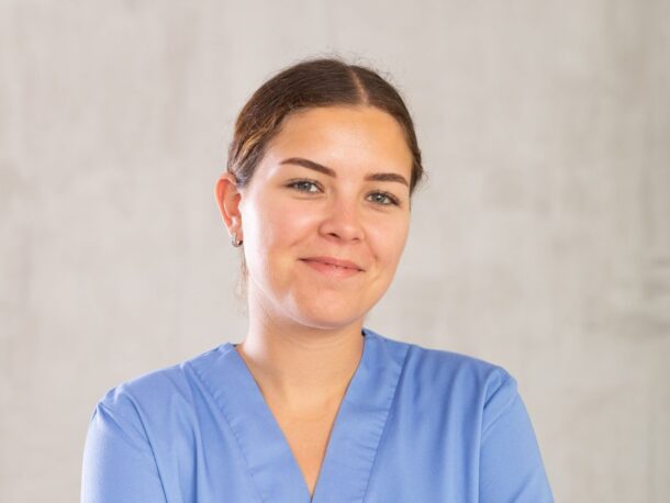 A young woman smiles at the camera. She's wearing blue scrubs and her brown hair is tied back. Her skin is lightly tanned and she has hazel eyes.