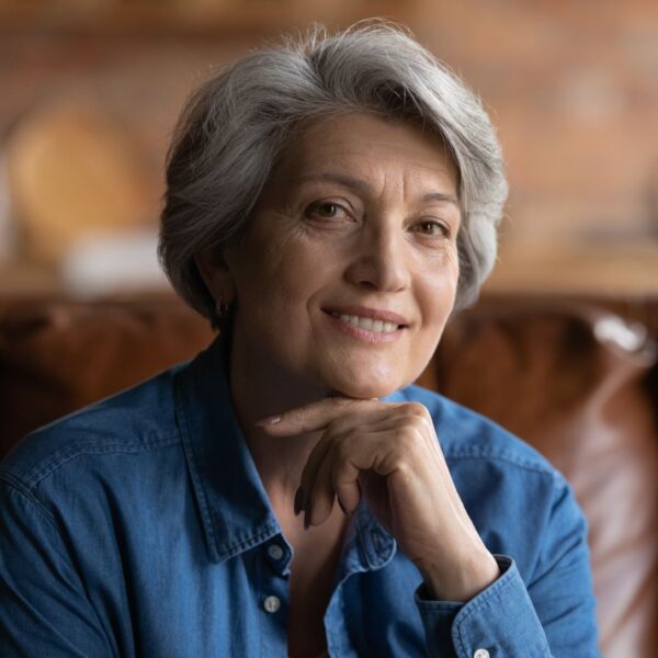 An older woman with short grey hair sits on a sofa and smiles as she rests her chin on her hand, she is wearing a dark denim shirt.