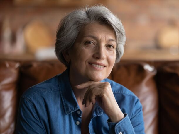 An older woman with short grey hair sits on a sofa and smiles as she rests her chin on her hand, she is wearing a dark denim shirt.