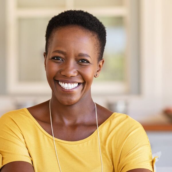 A woman with very short black coily hair smiles widely at the camera, she is wearing a yellow blouse.