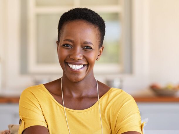 A woman with very short black coily hair smiles widely at the camera, she is wearing a yellow blouse.