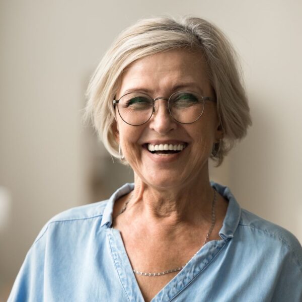 An older woman with short, greying blonde hair smiles widely at the camera, she wears round, wire framed glasses and a denim shirt.