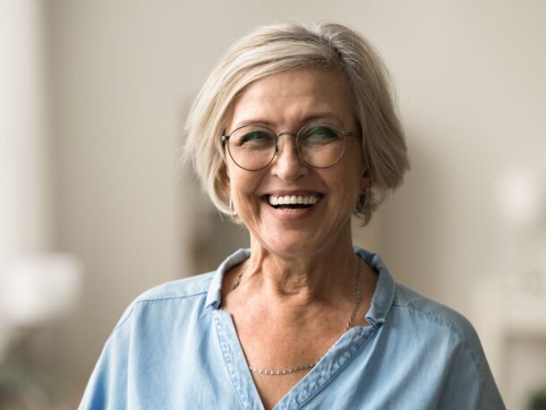An older woman with short, greying blonde hair smiles widely at the camera, she wears round, wire framed glasses and a denim shirt.