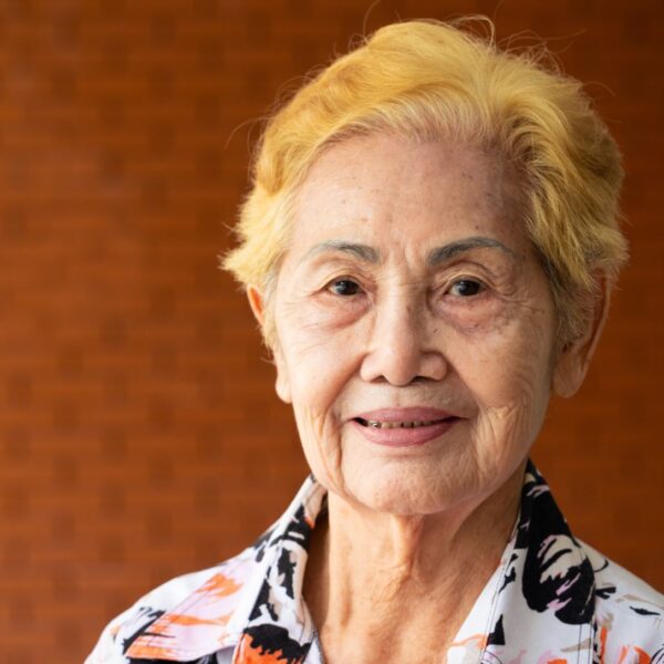 An older woman with dyed yellow hair smiles gently at the camera, she is standing in front of a red brick wall and wears a floral patterned shirt.
