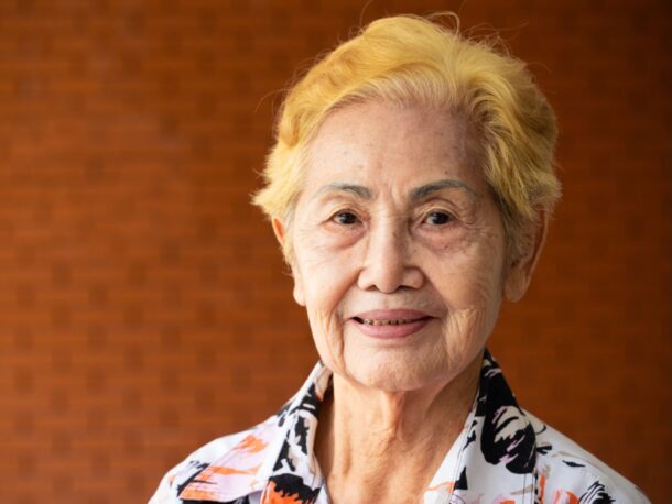 An older woman with dyed yellow hair smiles gently at the camera, she is standing in front of a red brick wall and wears a floral patterned shirt.