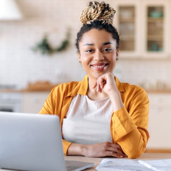 A woman sits at a desk with a laptop in front of her resting her chin on her hand and smiling at the camera.