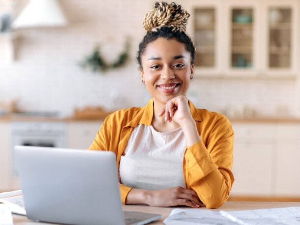 A woman sits at a desk with a laptop in front of her resting her chin on her hand and smiling at the camera.