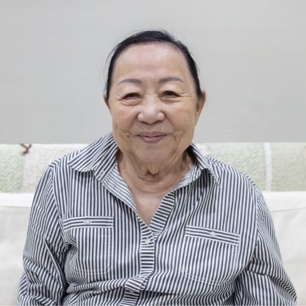 An older woman sits on a sofa smiling at the camera, she wears a white and black striped shirt.