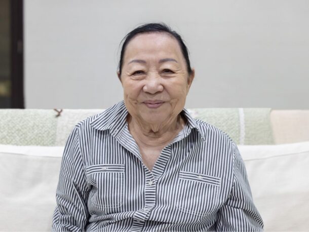 An older woman sits on a sofa smiling at the camera, she wears a white and black striped shirt.
