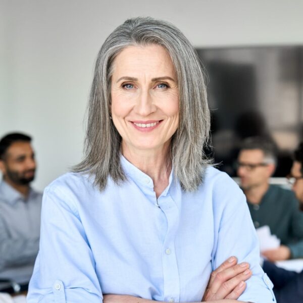 An older woman with shoulder length grey hair and blue eyes smiles at the camera, behind her people in business wear are having a discussion.