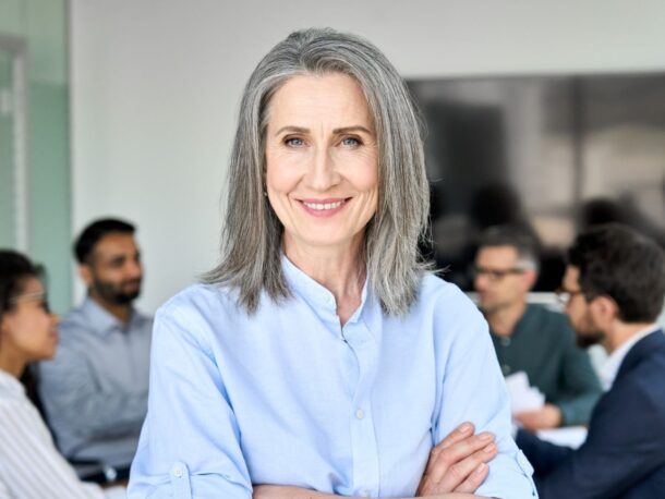 An older woman with shoulder length grey hair and blue eyes smiles at the camera, behind her people in business wear are having a discussion.
