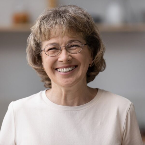 An older woman with short curly greying light brown hair smiles at the camera, she wears wire frame glasses and gold earrings.