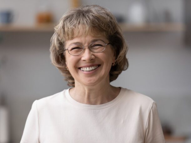 An older woman with short curly greying light brown hair smiles at the camera, she wears wire frame glasses and gold earrings.