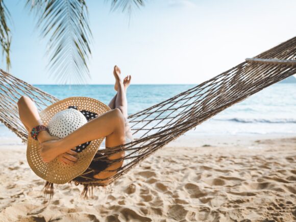 View of a woman sitting in a hammock on a tropical beach from behind, she wears a big sunhat and has her hands on her head, she is looking out to sea.