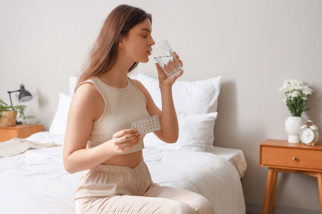A young woman sits on the edge of her bed, she is taking a sip of water from a glass and in the other hand holding a blisterpack of pink and white pills.