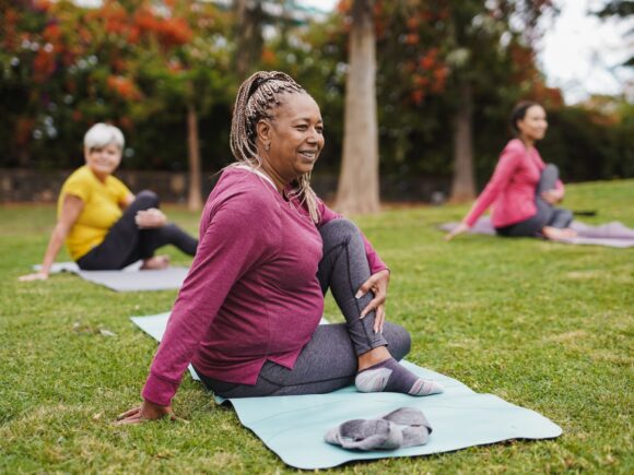 A group of three woman are doing yoga on mats in a park, the focus is on the woman centre front, they are in a seated pose.