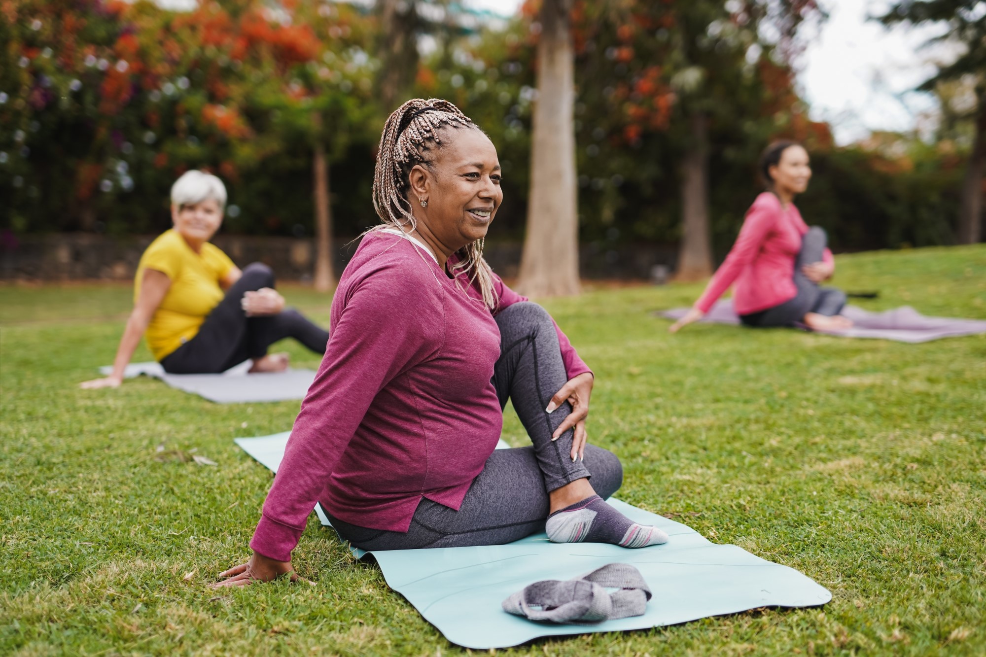 A group of three woman are doing yoga on mats in a park, the focus is on the woman centre front, they are in a seated pose.