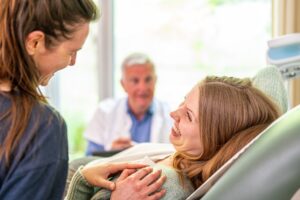 One woman reclines in a medical chair and smiles widely at another woman who places a hand on her shoulder. There is a healthcare professional in the background.