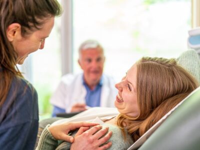 One woman reclines in a medical chair and smiles widely at another woman who places a hand on her shoulder. There is a healthcare professional in the background.