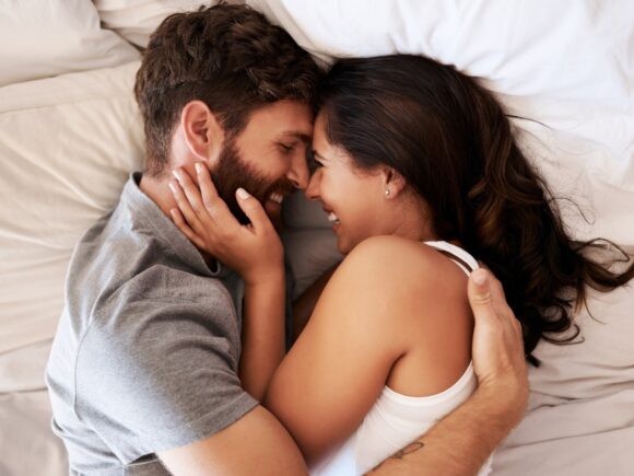 A young couple lie in bed together embracing and laughing, she cups his bearded chin as he places a hand on her back.