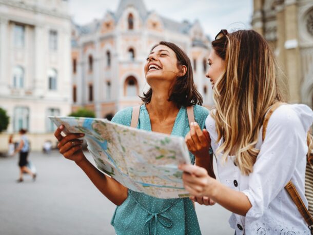 Two young women burst into laughter as they look at a map, they appear to be holidaying in a European city standing in a plaza outside a cathedral.