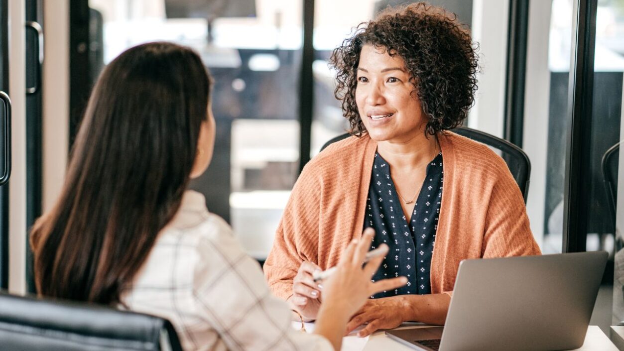 Two women talk to each other across a desk in an office, one has her back to the camera.