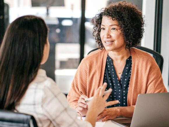 Two women talk to each other across a desk in an office, one has her back to the camera.