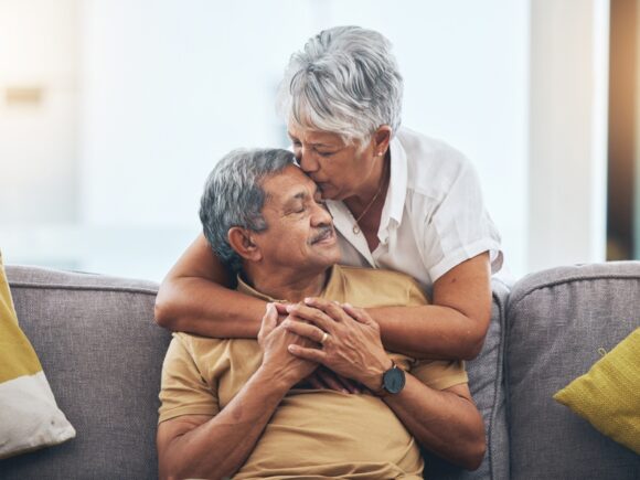 An older man sits on a sofa while his wife wraps her arms round him from behind and kisses him on the forehead.