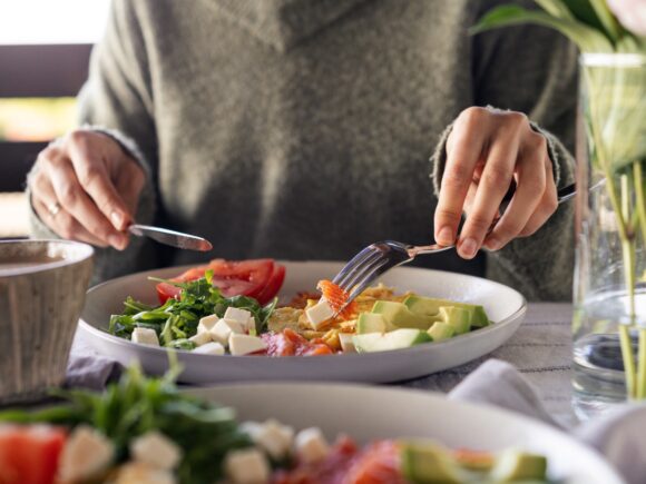 Close-up of a person cutting food on a plate with a knife and fork, the food looks healthy including avocado, tomatoes and spinach.