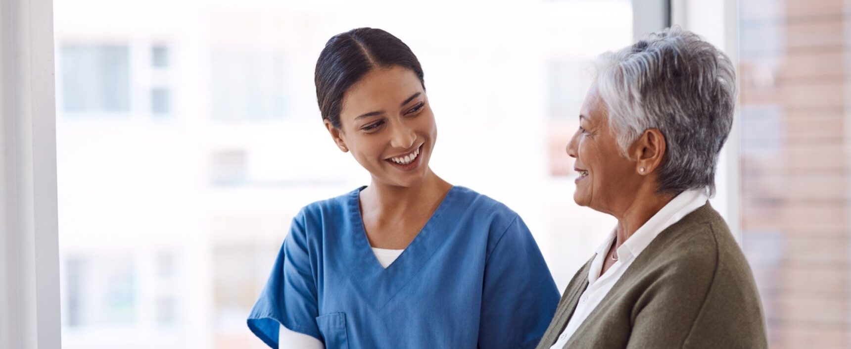 A healthcare professional holds an older woman's arm while they talk and and smile each other.