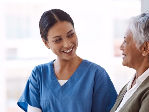 A healthcare professional holds an older woman's arm while they talk and and smile each other.