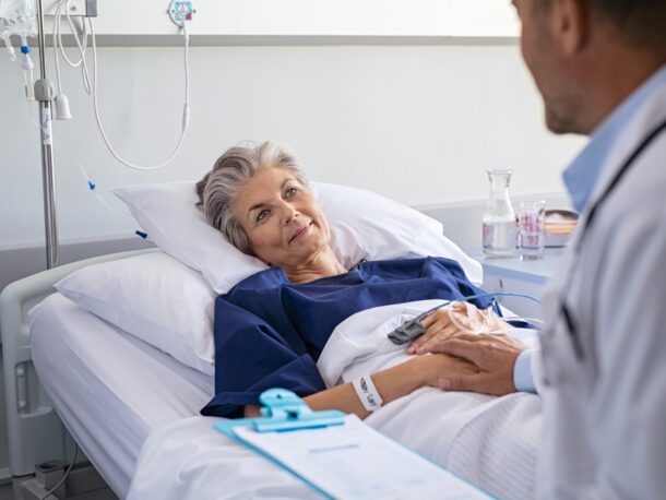 An older woman lies in a hospital bed and smiles up a doctor who is sat next to her holding her hand.