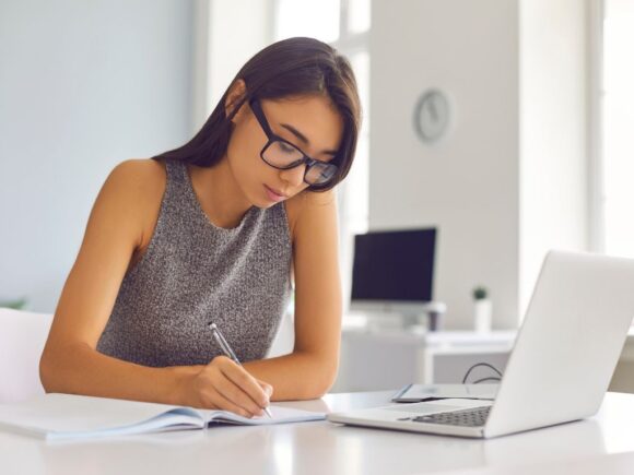 A woman wearing glasses writes in a notebook on a desk where there is also a laptop.
