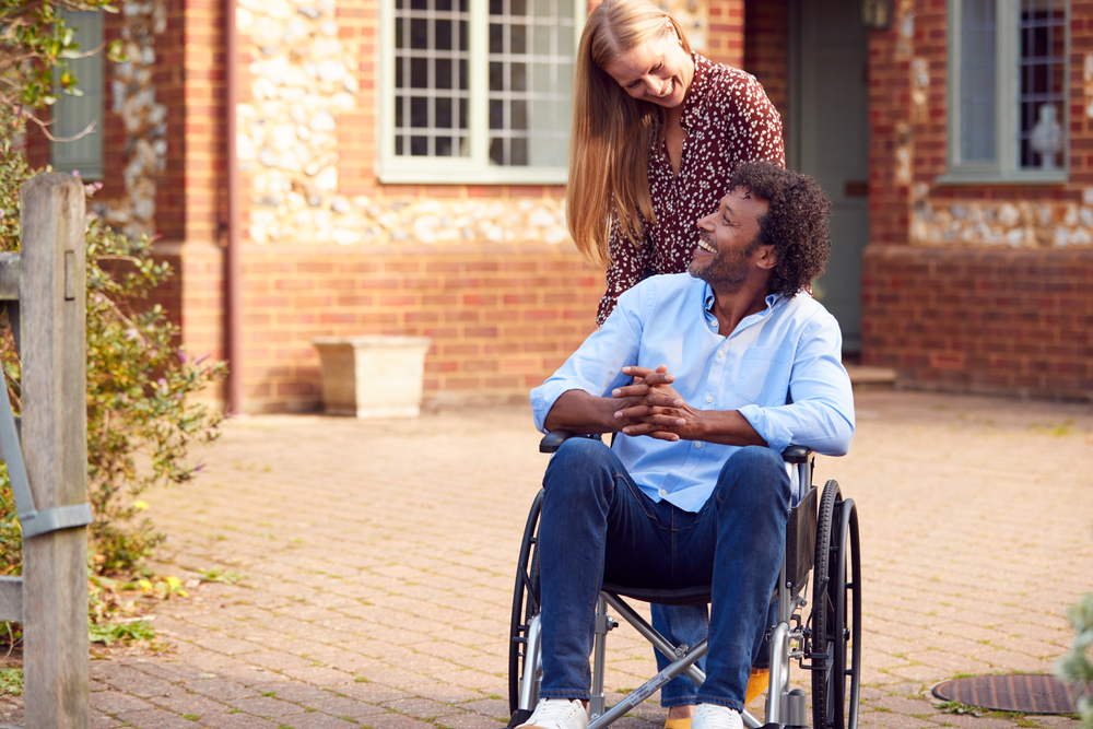 A man in a wheelchair smiles at a woman who is standing behind him.