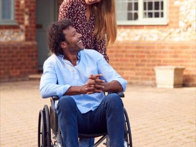 A man in a wheelchair smiles at a woman who is standing behind him.