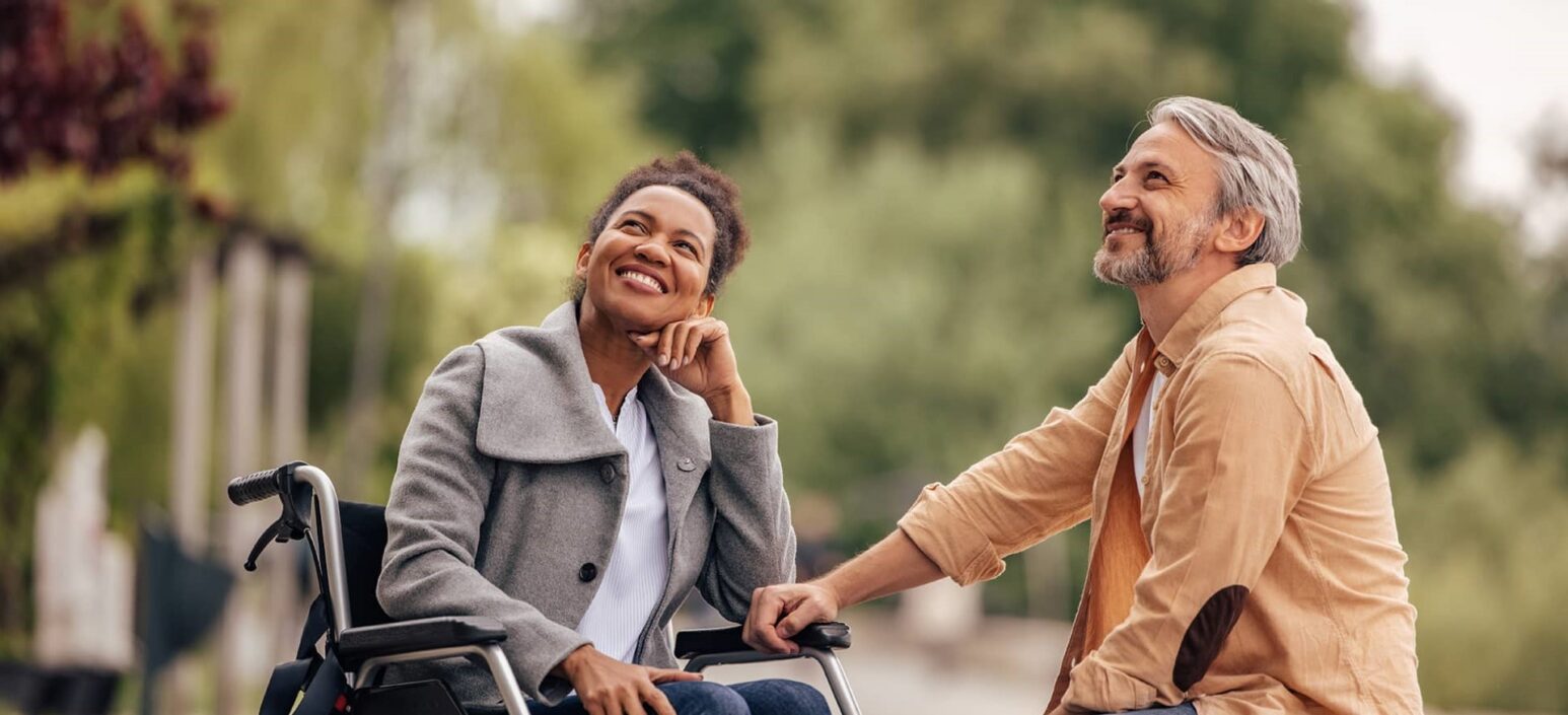 A woman in a wheelchair and a man sat in front of her look upwards smiling.