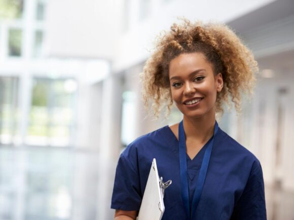 A young healthcare professional smiles at the camera while holding a clipboard.
