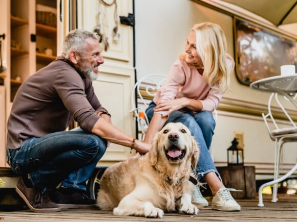A couple sit on a porch outside their caravan petting their pet golden retriever and smiling at each other.