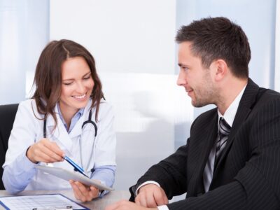 A doctor smiles and points at a tablet with a pen while a man in a suit smiles at her.